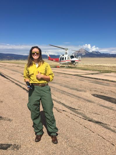 Diane wearing a yellow nomex shirt and green nomex fire pants gives a thumbs up standing in front of a plane