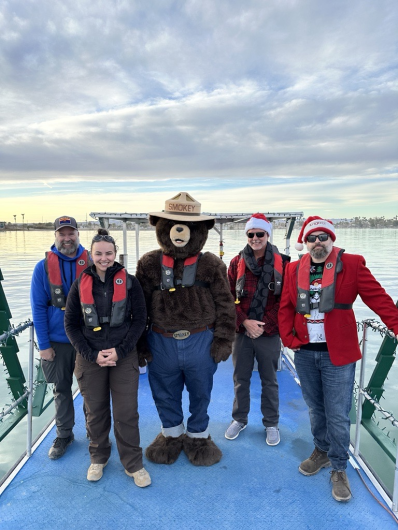 A group of people dressed up in cold weather gear stand on a boat with Smokey Bear