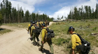 Caucasian men in yellow shirts, black packs, and black hardhats walk in single file on a dirt road.