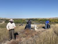 Photo. Daytime, sunny, clear-blue skies. Three people using rakes and black trash bags to clean up large piles of dried out weeds and brush. 